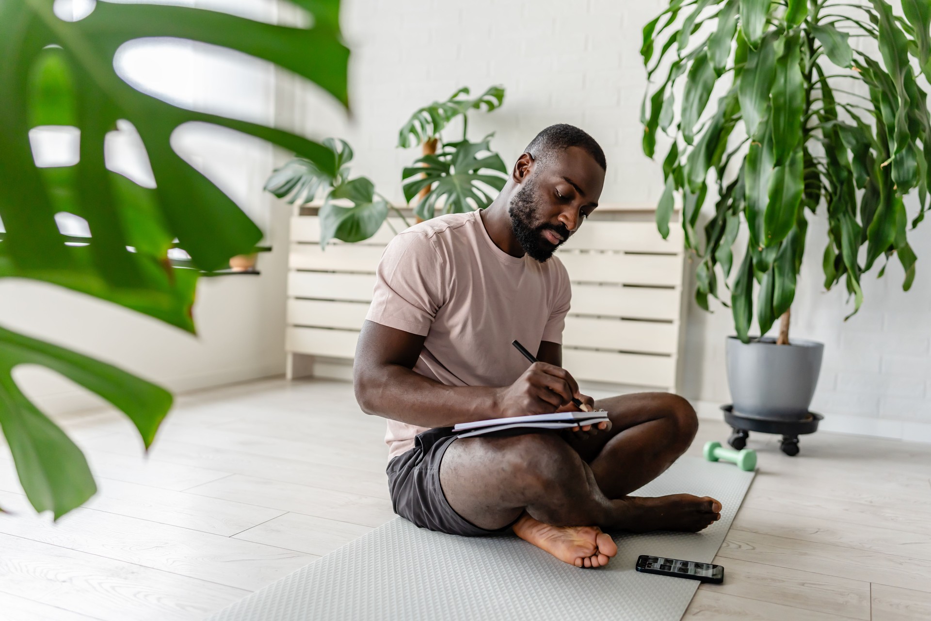 Young African-American man sitting cross-legged on a yoga mat at home, writing in a notebook after a workout, with a smartphone nearby, creating a calm and focused atmosphere.