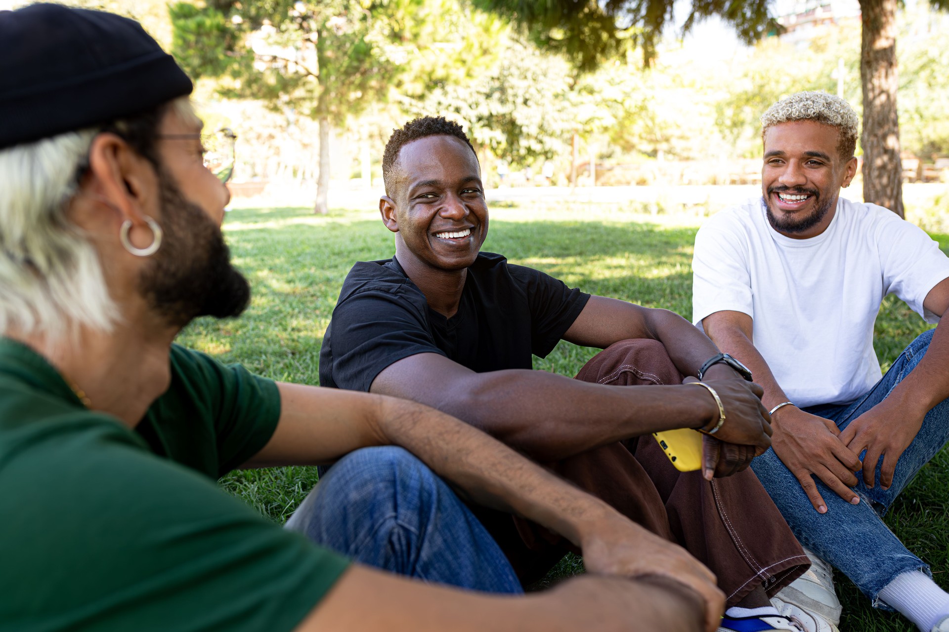 Young African American man and his male friends laughing and having fun together sitting on the grass in a park. Sharing thoughts, feelings, and struggles. 